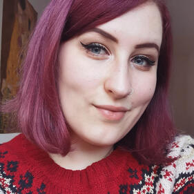 A young woman with shoulder-length, purple hair and winged eyeliner smiles at the camera.